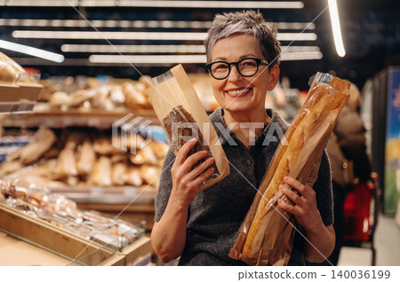 Middle-aged woman with short gray hair holding two loaves of bread in a bakery, shelves filled with various baked goods in the background 140036199