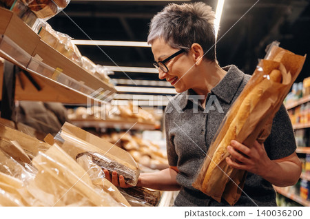 Middle-aged woman with short gray hair wearing glasses examines packaged bread while holding a baguette in a grocery store aisle filled with various baked goods Middle-aged woman with short gray hair wearing glasses examines packaged bread while holding a baguette in a grocery store aisle filled with various baked goods 140036200