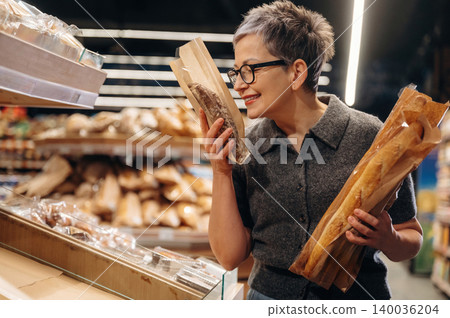 Middle-aged woman with short gray hair wearing glasses holds two baguettes while smelling one inside a bakery with various bread loaves displayed in the background Middle-aged woman with short gray hair wearing glasses holds two baguettes while smelling one inside a bakery with various bread loaves displayed in the background 140036204