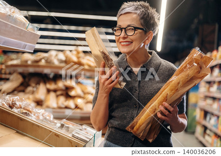 Mature woman with short gray hair wearing glasses holds fresh baguettes in a bakery aisle, surrounded by various types of bread on display in a grocery store Mature woman with short gray hair wearing glasses holds fresh baguettes in a bakery aisle, surrounded by various types of bread on display in a grocery store 140036206