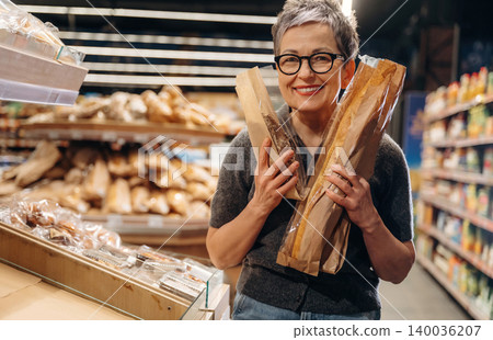 Middle-aged woman with short gray hair holding two baguettes in a grocery store aisle filled with bread and various products on shelves in the background Middle-aged woman with short gray hair holding two baguettes in a grocery store aisle filled with bread and various products on shelves in the background 140036207