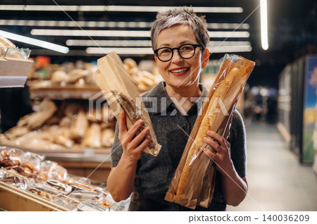 Middle-aged woman with short gray hair smiles while holding two loaves of bread in a bakery, with various baked goods displayed in the background Middle-aged woman with short gray hair smiles while holding two loaves of bread in a bakery, with various baked goods displayed in the background 140036209