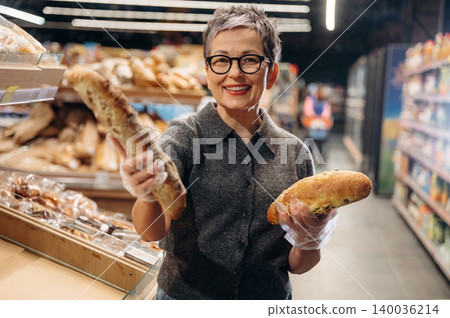 Middle-aged woman with short gray hair wearing glasses holds two types of bread in a bakery aisle, shelves filled with various baked goods in the background Middle-aged woman with short gray hair wearing glasses holds two types of bread in a bakery aisle, shelves filled with various baked goods in the background 140036214