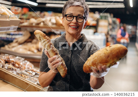 Mature woman with short gray hair wearing glasses holds two types of bread in a bakery, shelves of various baked goods visible in the background Mature woman with short gray hair wearing glasses holds two types of bread in a bakery, shelves of various baked goods visible in the background 140036215