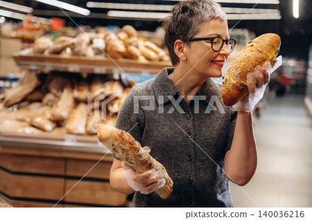 Woman with short gray hair wearing glasses holds fresh bread in a bakery, surrounded by various types of loaves on display in the background Woman with short gray hair wearing glasses holds fresh bread in a bakery, surrounded by various types of loaves on display in the background 140036216
