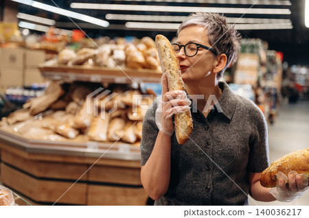 Mature woman with short gray hair wearing glasses holds a fresh baguette close to her face inside a bakery, shelves of bread visible in the background 140036217
