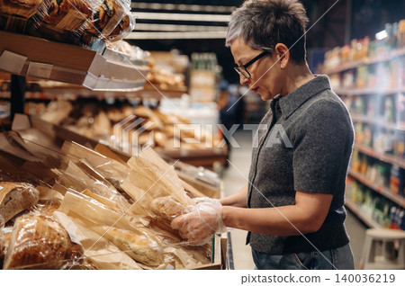 Middle-aged woman wearing glasses inspects packaged bread products on display in a grocery store aisle with shelves filled with various food items in the background Middle-aged woman wearing glasses inspects packaged bread products on display in a grocery store aisle with shelves filled with various food items in the background 140036219