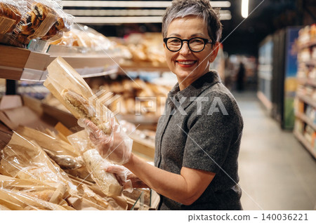 Mature woman with short gray hair wearing glasses and gloves holds a loaf of bread while shopping in a bakery section of a grocery store filled with baked goods Mature woman with short gray hair wearing glasses and gloves holds a loaf of bread while shopping in a bakery section of a grocery store filled with baked goods 140036221