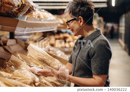 Mature woman with short hair wearing glasses inspects packaged bread in a bakery section of a grocery store, shelves filled with various baked goods in the background Mature woman with short hair wearing glasses inspects packaged bread in a bakery section of a grocery store, shelves filled with various baked goods in the background 140036222