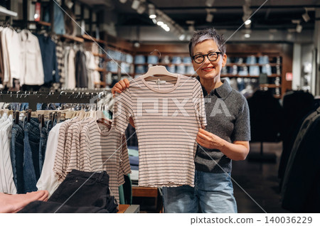 Asian female customer in gray shirt holds striped t-shirt while shopping in clothing store, racks of apparel and organized displays visible in the background Asian female customer in gray shirt holds striped t-shirt while shopping in clothing store, racks of apparel and organized displays visible in the background 140036229