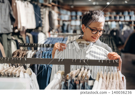 Middle-aged woman with short gray hair wearing glasses examines a striped shirt while shopping in a clothing store with various garments displayed in the background Middle-aged woman with short gray hair wearing glasses examines a striped shirt while shopping in a clothing store with various garments displayed in the background 140036236