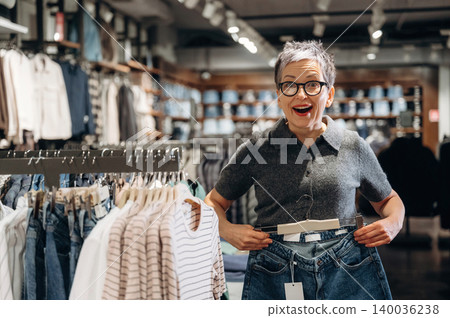 Mature woman with short gray hair smiles while holding a pair of jeans in a clothing store, surrounded by racks of apparel and a modern retail environment Mature woman with short gray hair smiles while holding a pair of jeans in a clothing store, surrounded by racks of apparel and a modern retail environment 140036238