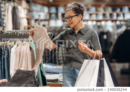 Asian woman shopping in a clothing store, examining a striped shirt while holding shopping bags, with racks of apparel and a wooden display in the background Asian woman shopping in a clothing store, examining a striped shirt while holding shopping bags, with racks of apparel and a wooden display in the background 140036244