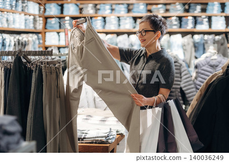 Woman examining beige trousers while shopping in a clothing store, surrounded by racks of apparel and neatly arranged garments in the background 140036249