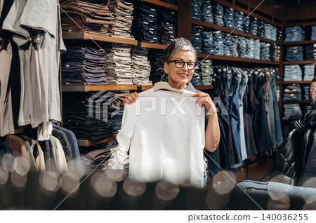Middle-aged woman with short gray hair holds a white sweater while standing in a clothing store, shelves filled with folded garments in the background 140036255