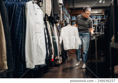 Senior woman shopping for clothing in a retail store, holding a white sweatshirt while browsing through racks of apparel and denim jeans in the background 140036257