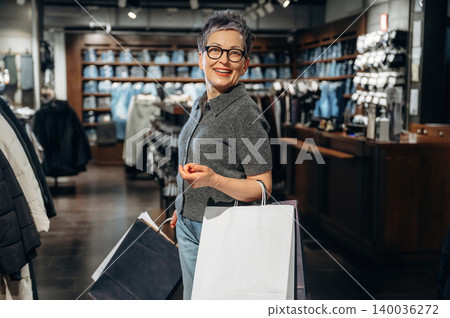 Mature woman with short gray hair wearing glasses smiles while holding shopping bags inside a modern clothing store with racks of apparel in the background 140036272