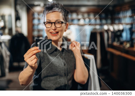 Mature woman with short gray hair and glasses holds a black credit card while smiling, standing in a clothing store with racks of apparel in the background Mature woman with short gray hair and glasses holds a black credit card while smiling, standing in a clothing store with racks of apparel in the background 140036300