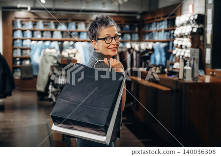 Mature woman with short gray hair and glasses smiles while holding shopping bags inside a clothing store with neatly arranged apparel in the background 140036308