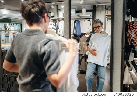 Woman with short hair wearing glasses examines white shirt in front of mirror inside clothing store, casual attire and fashion items displayed in the background Woman with short hair wearing glasses examines white shirt in front of mirror inside clothing store, casual attire and fashion items displayed in the background 140036320