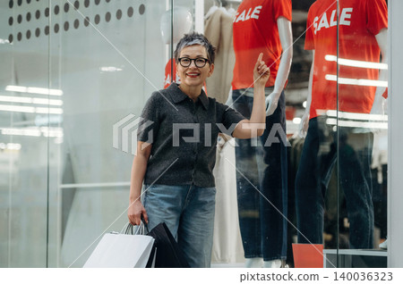 Senior Asian woman with glasses holding shopping bags stands in front of store window displaying red SALE shirts and mannequins, smiling and pointing at the sale sign Senior Asian woman with glasses holding shopping bags stands in front of store window displaying red SALE shirts and mannequins, smiling and pointing at the sale sign 140036323