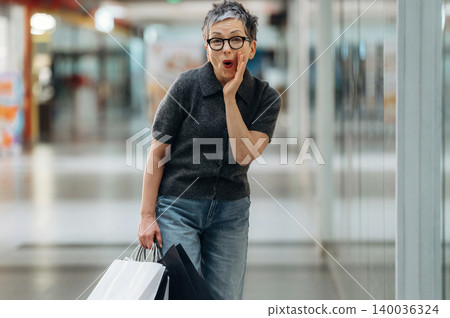 Senior woman with short gray hair wearing glasses holds shopping bags and poses playfully in a modern shopping mall with reflective surfaces and bright lighting Senior woman with short gray hair wearing glasses holds shopping bags and poses playfully in a modern shopping mall with reflective surfaces and bright lighting 140036324
