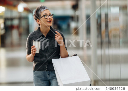Middle-aged woman with short gray hair holds coffee cup and shopping bag while walking through a modern shopping mall with bright lighting and glass walls Middle-aged woman with short gray hair holds coffee cup and shopping bag while walking through a modern shopping mall with bright lighting and glass walls 140036326
