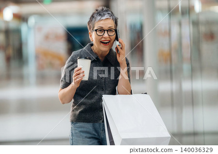 Senior woman with short gray hair wearing glasses talks on the phone while holding a coffee cup and shopping bag inside a modern shopping mall Senior woman with short gray hair wearing glasses talks on the phone while holding a coffee cup and shopping bag inside a modern shopping mall 140036329