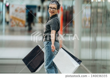 Mature woman with short gray hair smiles while holding shopping bags in a modern shopping mall, with blurred storefronts and reflective surfaces in the background Mature woman with short gray hair smiles while holding shopping bags in a modern shopping mall, with blurred storefronts and reflective surfaces in the background 140036335