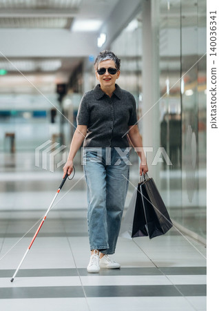 Elderly Asian woman wearing sunglasses walks confidently with a white cane and shopping bags in a modern shopping mall corridor with glass walls and bright lighting Elderly Asian woman wearing sunglasses walks confidently with a white cane and shopping bags in a modern shopping mall corridor with glass walls and bright lighting 140036341