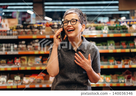 Middle-aged woman with short gray hair wearing glasses talks on a smartphone while smiling inside a grocery store with shelves of products in the background Middle-aged woman with short gray hair wearing glasses talks on a smartphone while smiling inside a grocery store with shelves of products in the background 140036346