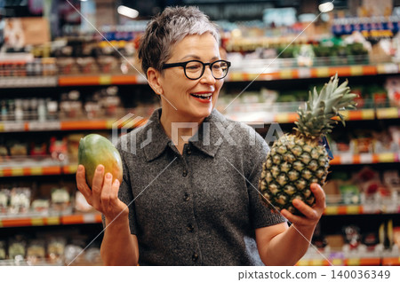 Mature woman with short gray hair wearing glasses holds a pineapple in one hand and a mango in the other inside a grocery store with shelves of products behind her Mature woman with short gray hair wearing glasses holds a pineapple in one hand and a mango in the other inside a grocery store with shelves of products behind her 140036349