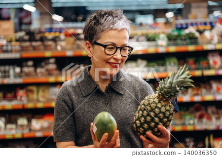 Mature woman with short gray hair wearing glasses holds pineapple and avocado in a grocery store aisle filled with various food products and shelves in the background Mature woman with short gray hair wearing glasses holds pineapple and avocado in a grocery store aisle filled with various food products and shelves in the background 140036350