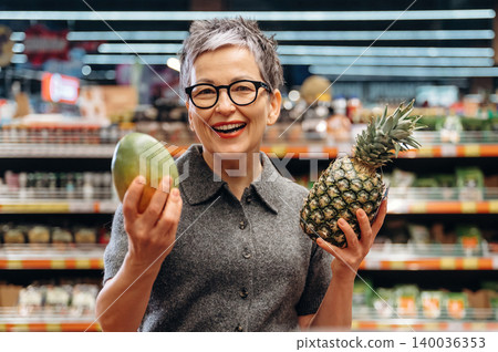 Mature woman with short gray hair wearing glasses holds a mango and pineapple while smiling in a grocery store aisle filled with fresh produce and colorful packaging Mature woman with short gray hair wearing glasses holds a mango and pineapple while smiling in a grocery store aisle filled with fresh produce and colorful packaging 140036353