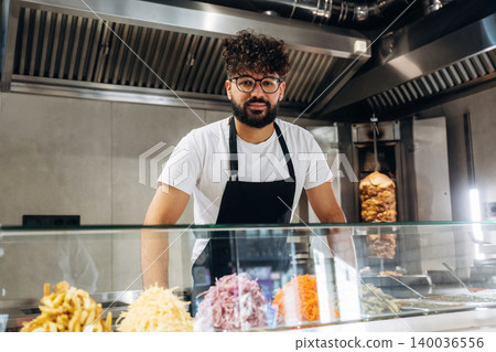Male chef with curly hair wearing glasses and black apron stands behind a glass counter displaying various fresh ingredients in a modern fast food restaurant 140036556