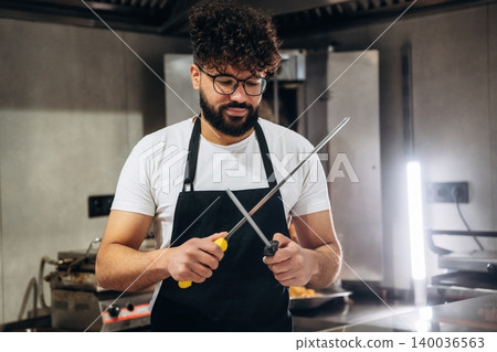 Male chef with curly hair wearing glasses sharpens kitchen knife with a honing steel in a modern restaurant kitchen with stainless steel appliances Male chef with curly hair wearing glasses sharpens kitchen knife with a honing steel in a modern restaurant kitchen with stainless steel appliances 140036563