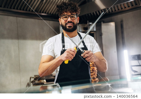 Male chef with curly hair wearing glasses holds kitchen knives in a modern kitchen, showcasing culinary tools and equipment in a professional cooking environment Male chef with curly hair wearing glasses holds kitchen knives in a modern kitchen, showcasing culinary tools and equipment in a professional cooking environment 140036564
