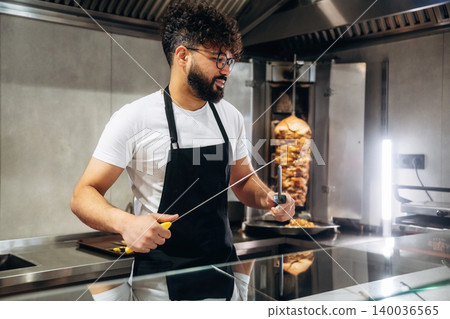 Male chef with curly hair wearing glasses and black apron holds a knife while preparing food in a modern kitchen with a vertical rotisserie in the background 140036565