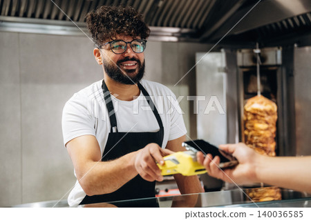Male cashier with curly hair wearing glasses hands over a payment terminal to a customer at a food counter with grilled meat in the background Male cashier with curly hair wearing glasses hands over a payment terminal to a customer at a food counter with grilled meat in the background 140036585