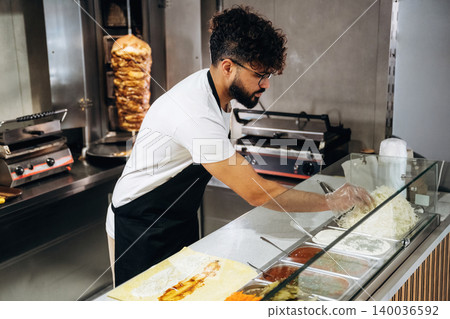 Male chef with curly hair prepares food in a restaurant kitchen, arranging rice and sauces in front of a vertical grill with meat in the background Male chef with curly hair prepares food in a restaurant kitchen, arranging rice and sauces in front of a vertical grill with meat in the background 140036592