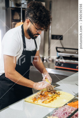 Male chef with curly hair prepares a wrap filled with vegetables and meat on a countertop in a modern kitchen with cooking equipment in the background Male chef with curly hair prepares a wrap filled with vegetables and meat on a countertop in a modern kitchen with cooking equipment in the background 140036598