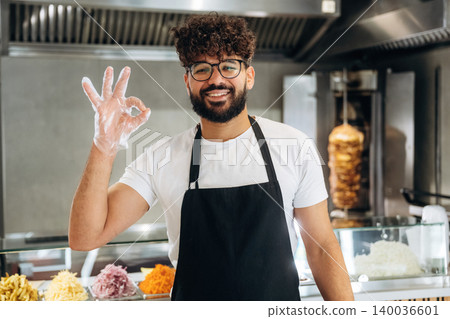 Male chef wearing glasses and gloves shows an okay hand gesture while standing in front of a food counter with various ingredients in a restaurant kitchen 140036601