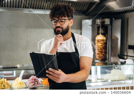Male restaurant worker with curly hair and glasses checks inventory on a tablet while standing in front of a display of fresh ingredients in a modern kitchen Male restaurant worker with curly hair and glasses checks inventory on a tablet while standing in front of a display of fresh ingredients in a modern kitchen 140036604