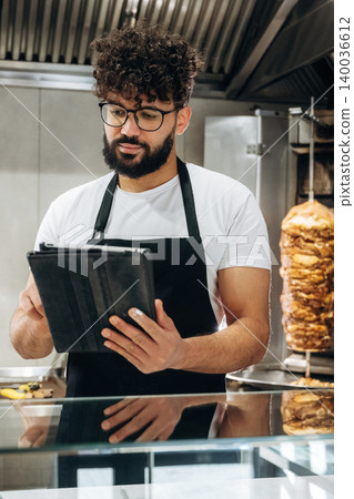 Male chef with curly hair wearing glasses and black apron checks orders on tablet in a restaurant kitchen with meat rotisserie in the background Male chef with curly hair wearing glasses and black apron checks orders on tablet in a restaurant kitchen with meat rotisserie in the background 140036612