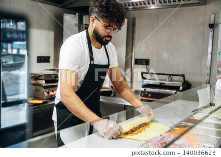 Male chef with curly hair wearing glasses prepares a wrap with meat and vegetables at a food counter in a modern restaurant kitchen Male chef with curly hair wearing glasses prepares a wrap with meat and vegetables at a food counter in a modern restaurant kitchen 140036623