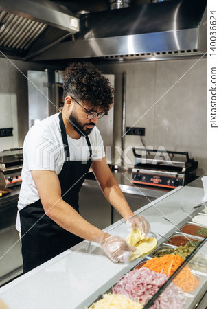 Male chef with curly hair wearing glasses prepares fresh ingredients in a modern kitchen, arranging vegetables and sauces on a countertop for meal preparation Male chef with curly hair wearing glasses prepares fresh ingredients in a modern kitchen, arranging vegetables and sauces on a countertop for meal preparation 140036624