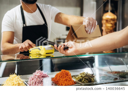 Male cashier in black apron processes payment with smartphone at food counter displaying colorful toppings in a fast-casual restaurant setting Male cashier in black apron processes payment with smartphone at food counter displaying colorful toppings in a fast-casual restaurant setting 140036645