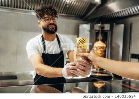 Male food vendor in black apron hands over wrapped kebab to customer at counter in modern fast food restaurant with grill in the background Male food vendor in black apron hands over wrapped kebab to customer at counter in modern fast food restaurant with grill in the background 140036654