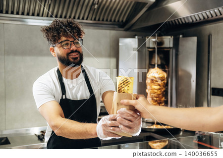 Male food vendor in black apron hands over wrapped kebab to customer at counter in modern fast-food restaurant with vertical grill in background 140036655