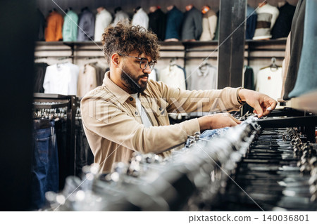 Male customer with curly hair browses clothing racks in a modern retail store, displaying various garments and organized shelves in the background Male customer with curly hair browses clothing racks in a modern retail store, displaying various garments and organized shelves in the background 140036801